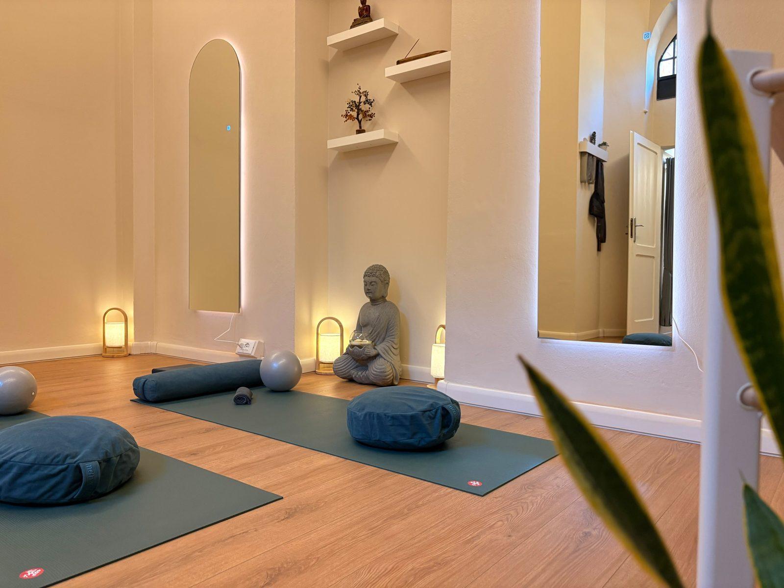Meditation cushions and bolsters on the mats, the Buddha statue between glowing lanterns, seen past a snake plant